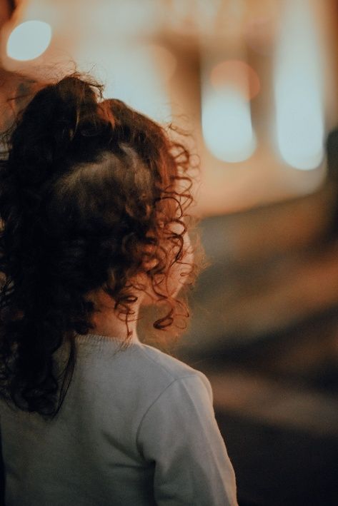 Person with curly hair in a white top, seen from behind, standing in warm blurred evening light.