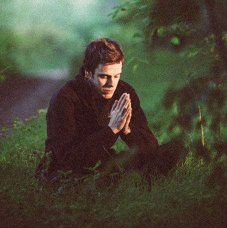 Man kneeling in a grassy field with hands clasped, bathed in green light.