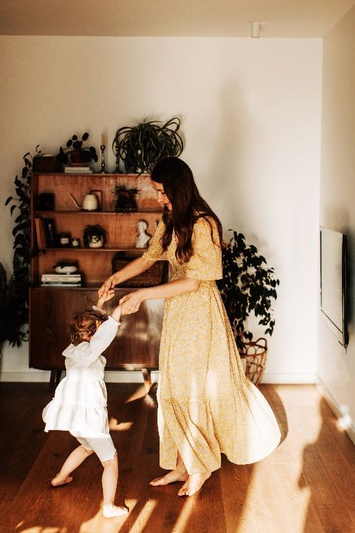 Woman dancing with a child in a sunlit living room with wooden shelves and plants