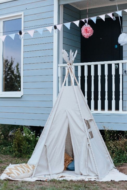 White teepee tent on a porch with bunting and pom-pom decorations, beside a light blue house