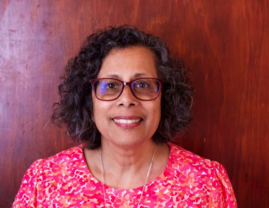 Smiling person with curly hair and glasses wearing a pink patterned top against a wooden background