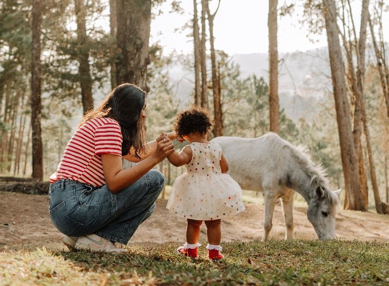 Woman and child pet a white horse in a wooded park, with a red striped shirt and white dress.