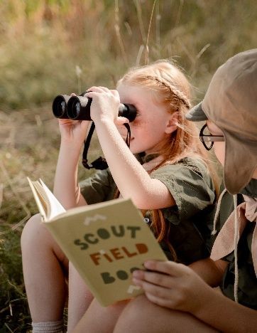Two scouts in green uniforms, one using binoculars while the other reads a Scout Field Book outdoors
