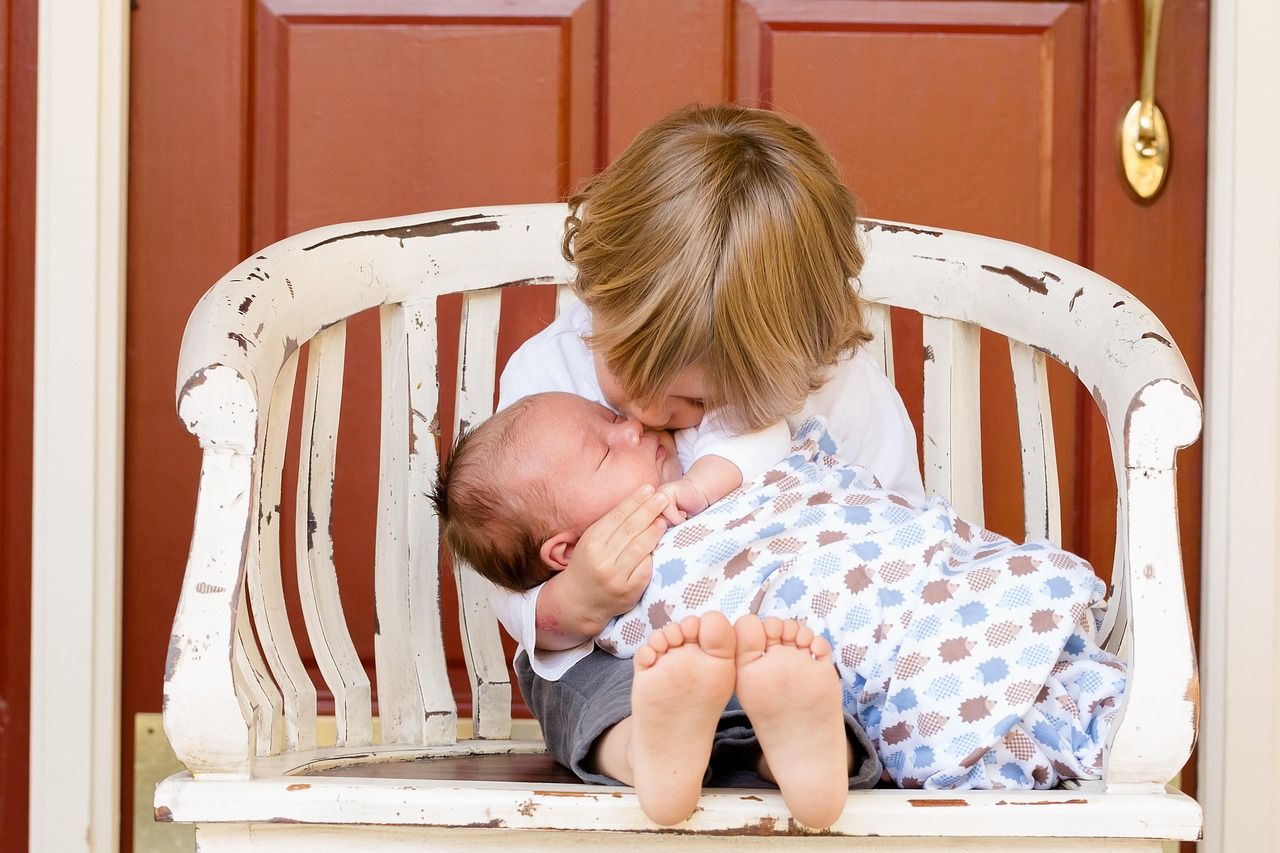 Older child kisses a baby, both on a weathered white bench in front of a red door.