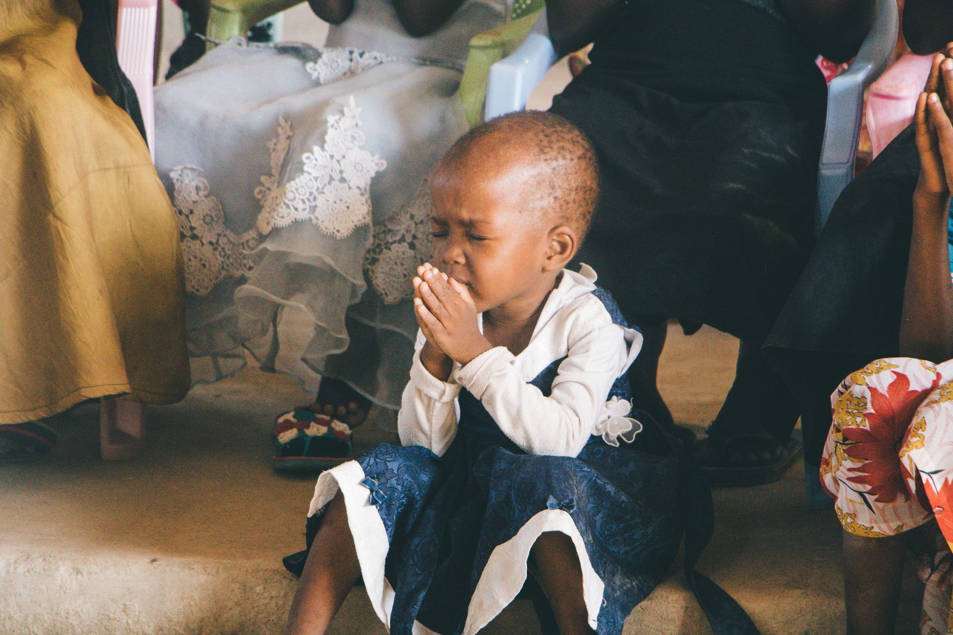 A person with eyes closed and hands clasped together in a prayer-like gesture, seated among others in a gathering.