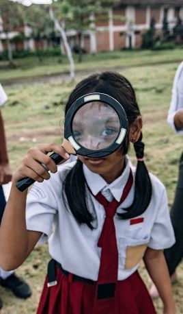 Student in a white school uniform and red tie holding a magnifying glass outdoors, looking closely at the camera.