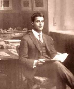 Seated man in a suit holding a book in a sepia-toned room with desk and papers.