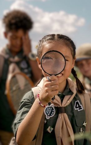 Girl in green scout uniform holding a magnifying glass up to her eye outdoors, with another scout behind her