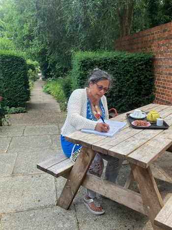 Person writing at a picnic table on a garden path, with food beside them.