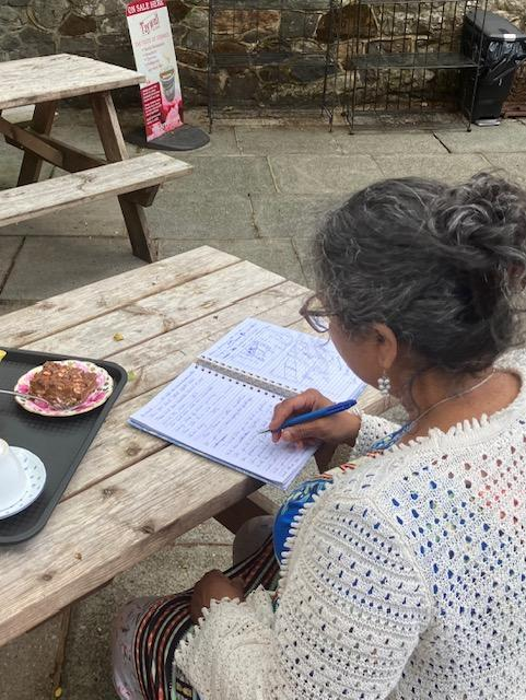 Person writing in a notebook at a wooden picnic table outdoors, with a small plate of food nearby.