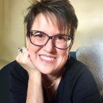 A woman wearing glasses is smiling with her hand on her chin.
