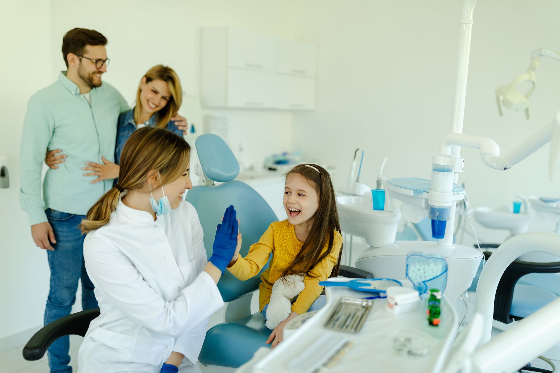 The dentist high-fives the young girl in the dental chair as her parents look on, smiling. The dentist high-fives the young girl in the dental chair as her parents look on, smiling.