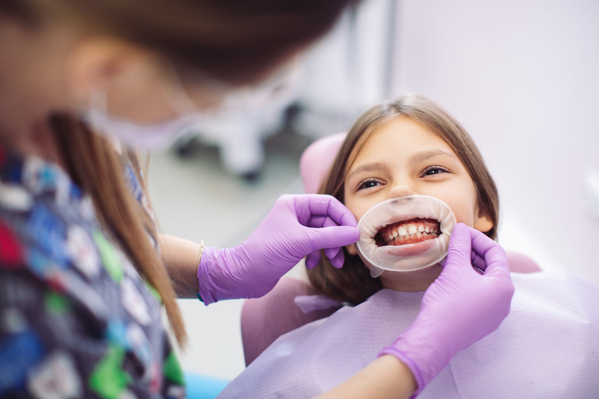 Dentist placing a cheek retractor in a patient's mouth at a dental clinic. Patient smiling, purple gloves.