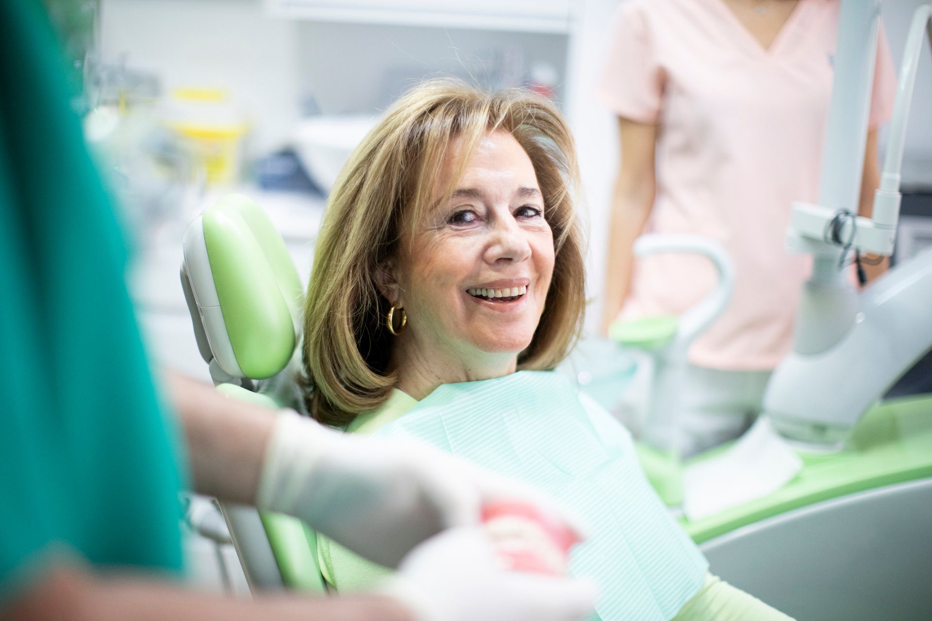 Woman smiles in a dental chair; dentist in green scrubs and dental assistant in pink shirt are present.