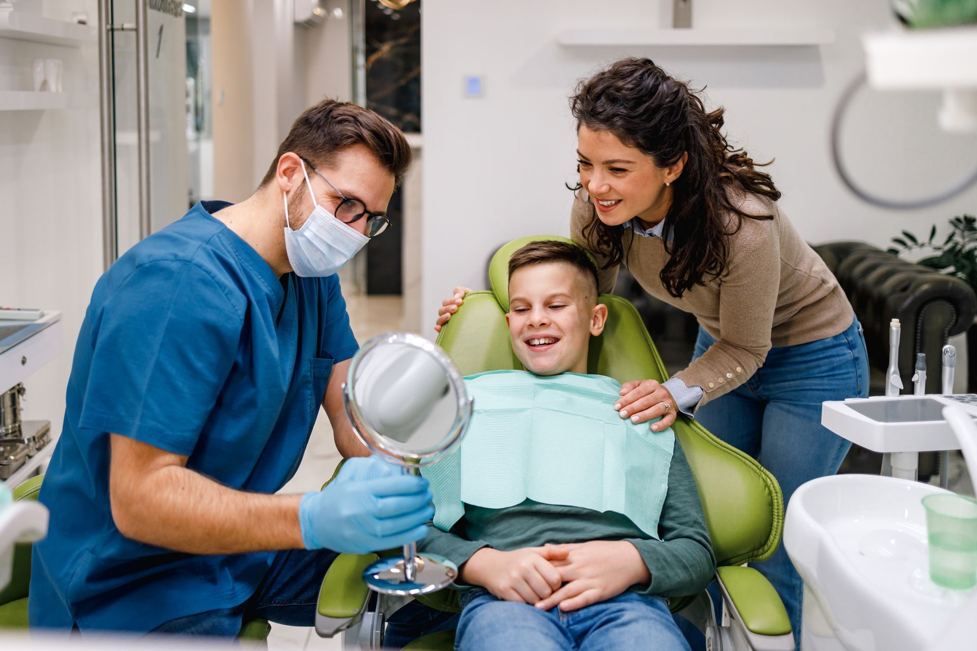 Dentist showing a young patient teeth in a mirror, mother watching. Dental office setting.