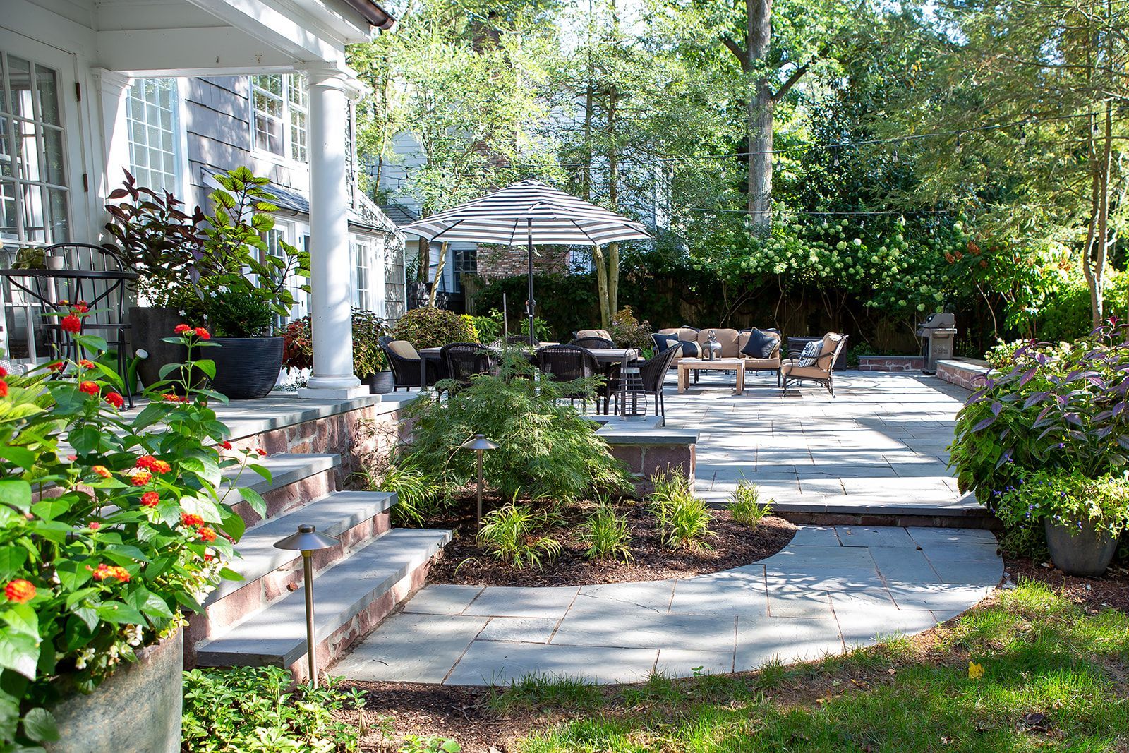 Stone patio with outdoor furniture, surrounded by greenery, next to a house with a porch.