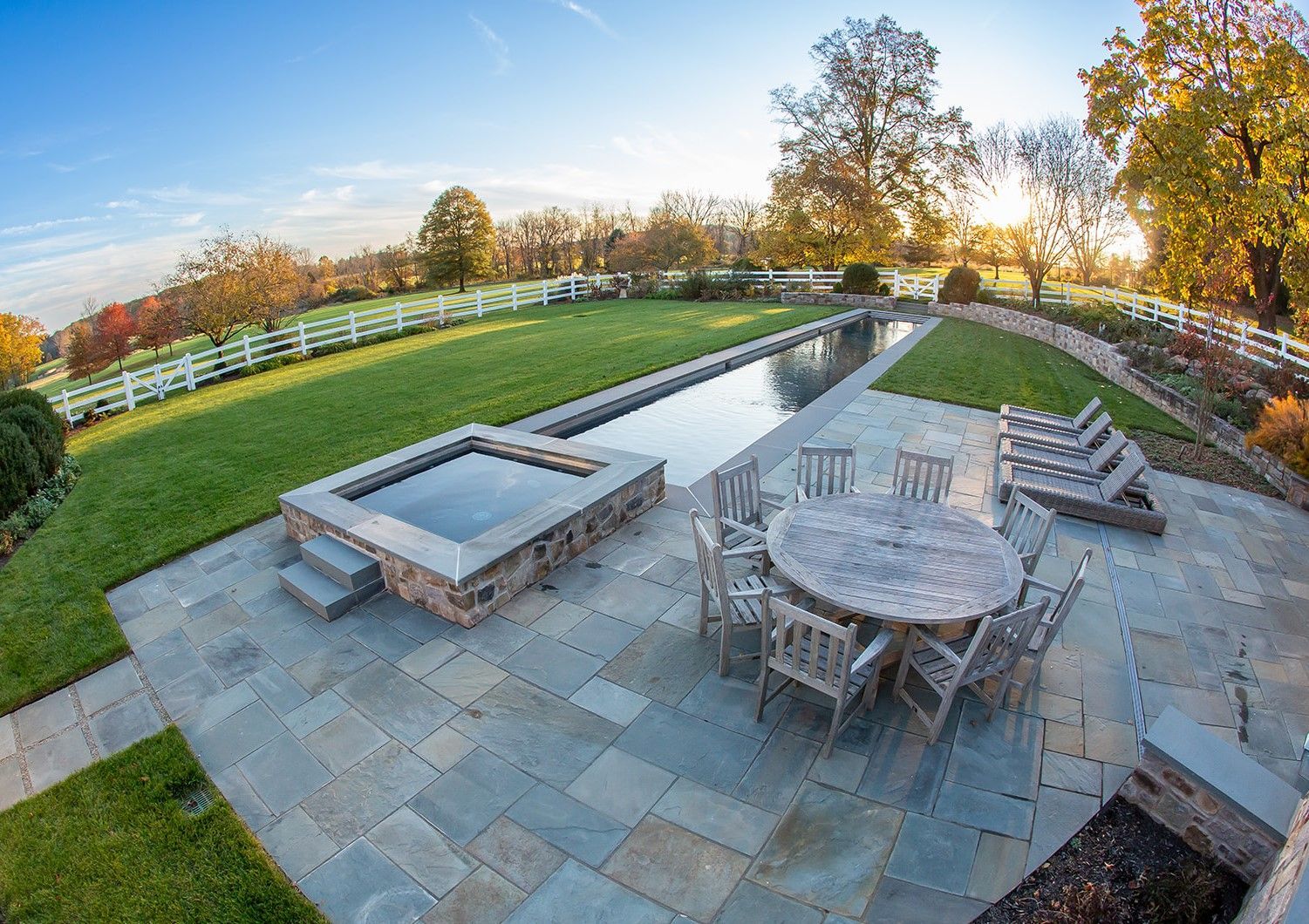 Stone patio with pool, hot tub, and round table set in a grassy yard with a white fence and trees.
