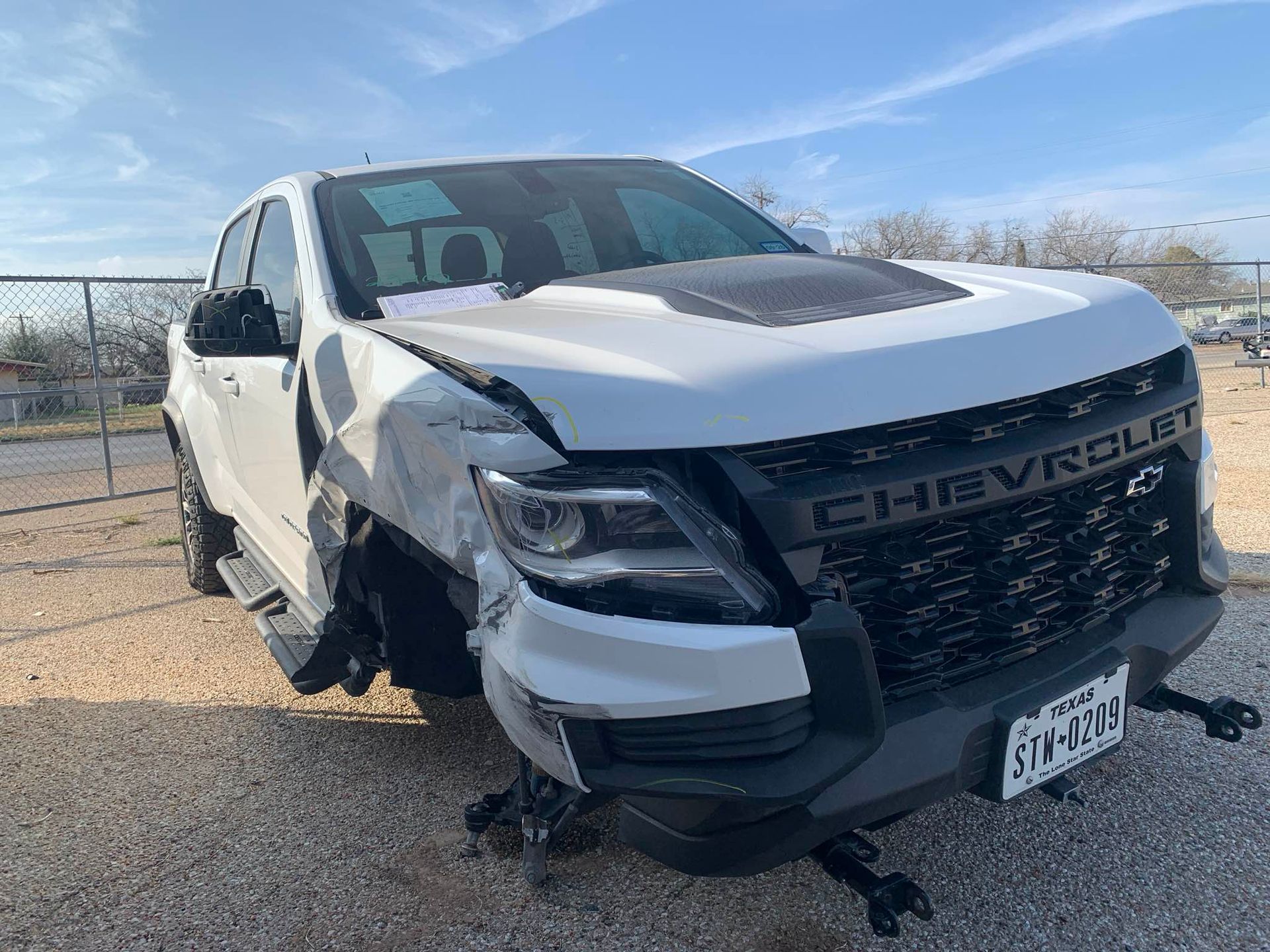 Damaged white Chevrolet pickup truck with front-end damage, parked outdoors on gravel near water.