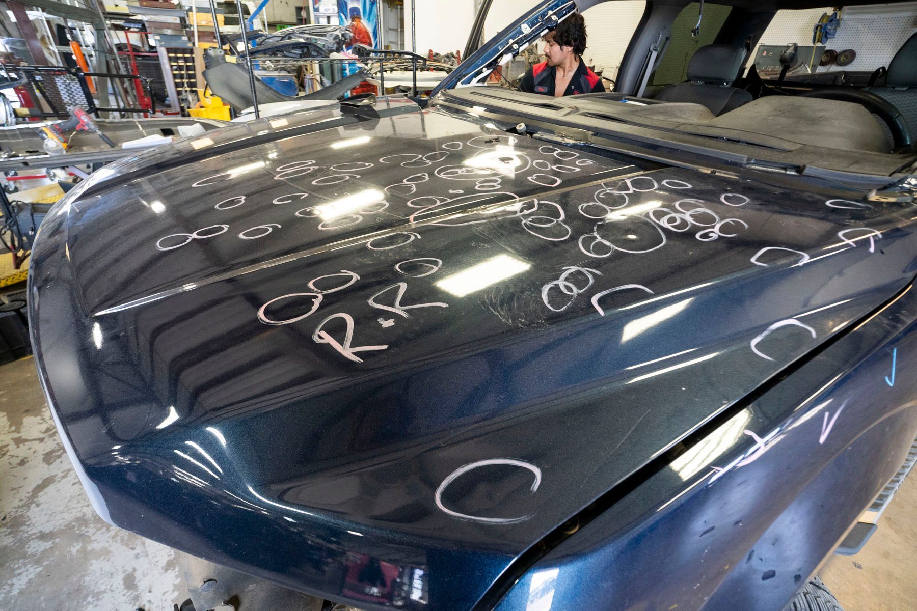 Dark blue car hood in an auto shop, covered with white marker notes and inspection circles