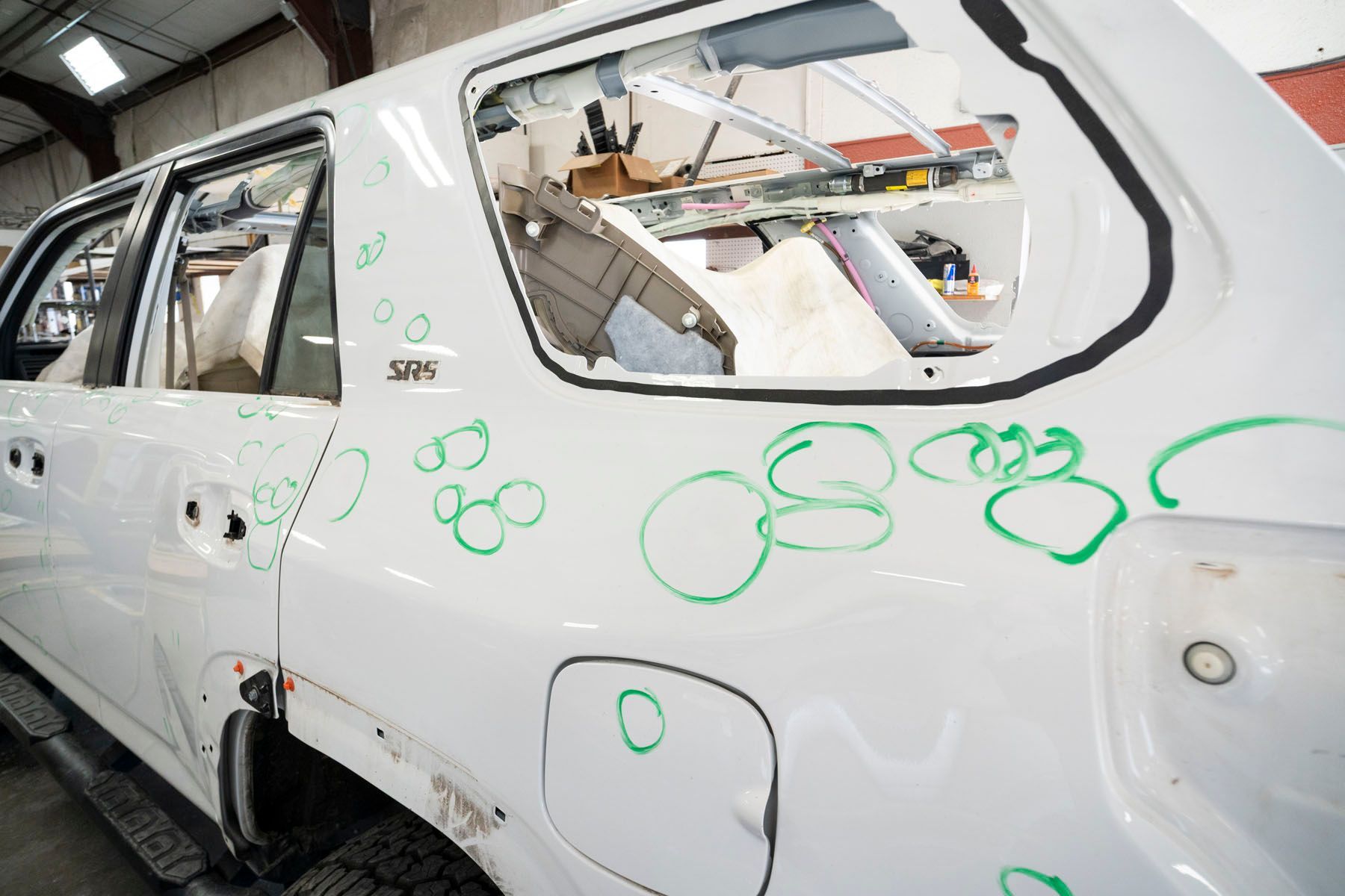 White car body in a repair shop, marked with green circles and showing damaged side panels and windows.