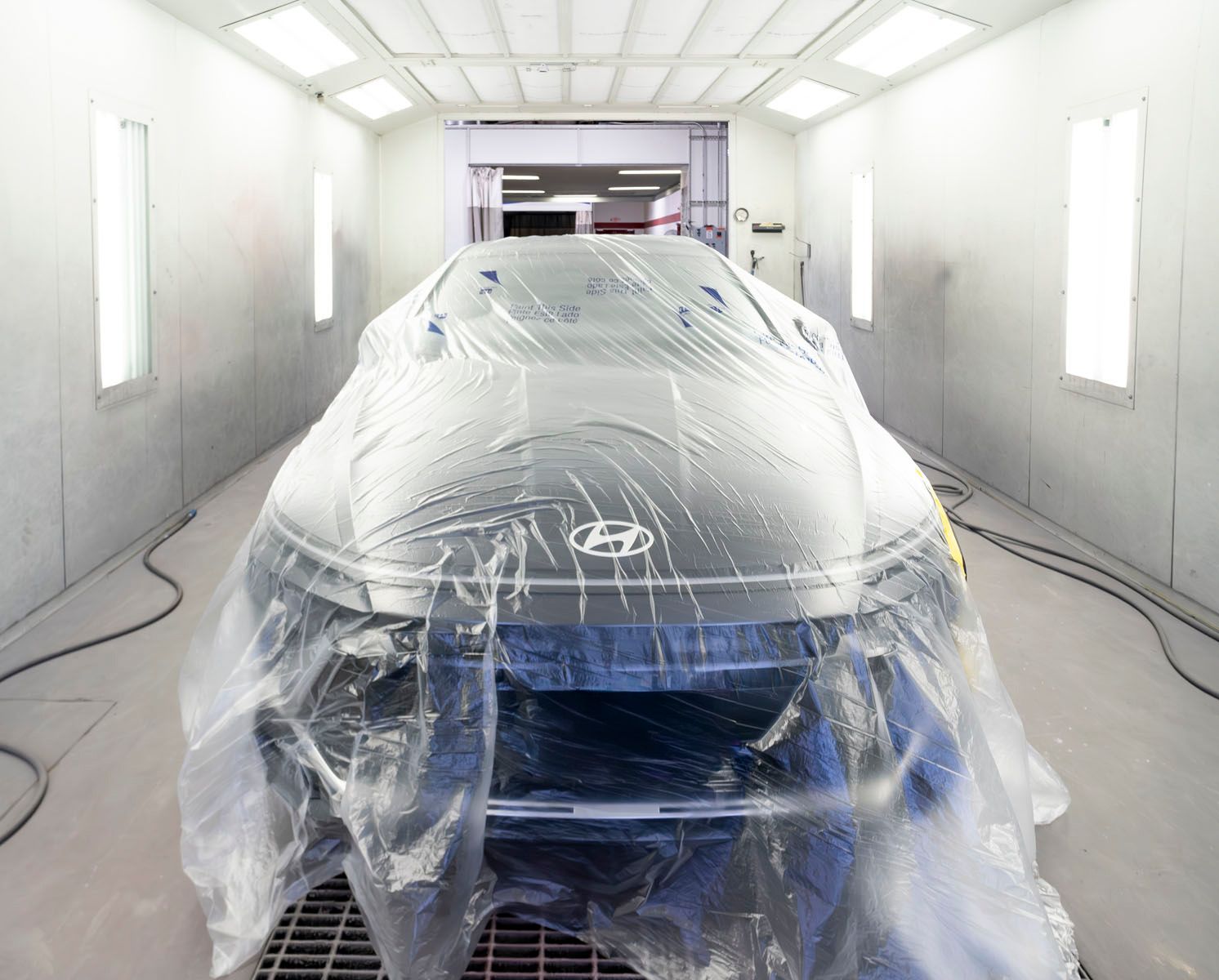 Car wrapped in plastic inside a white auto paint booth with bright overhead lights