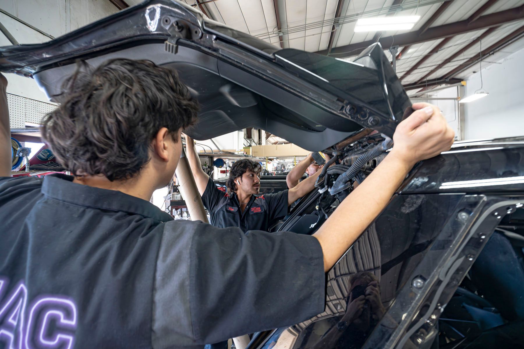 Two people work under an open car hood inside a garage.