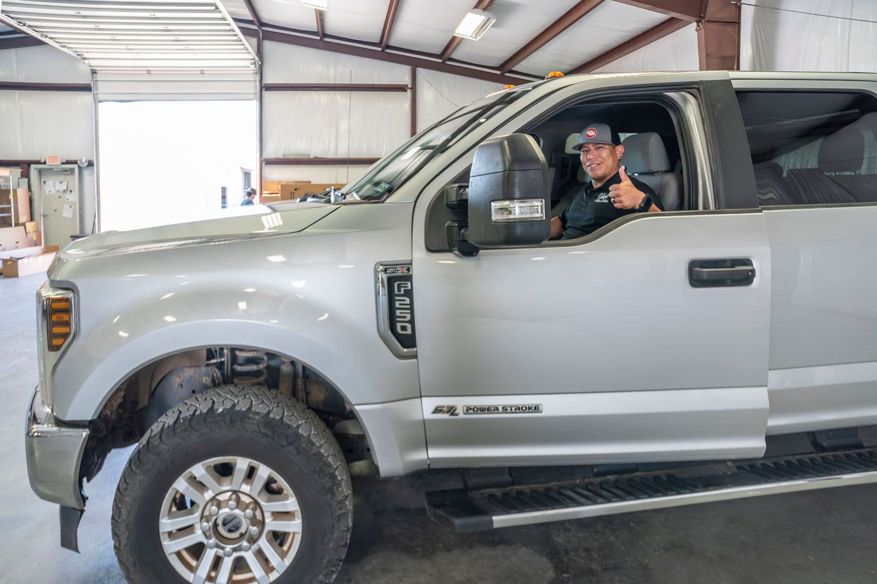 Man in a silver pickup truck inside a garage, smiling and waving from the driver’s seat