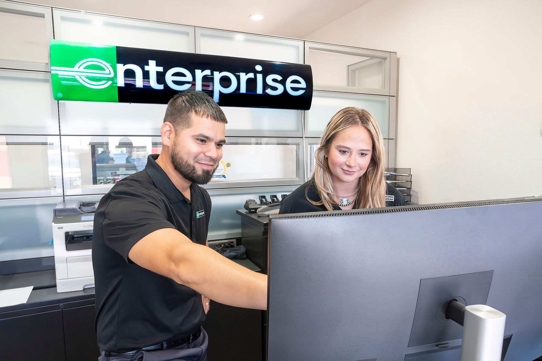 Two employees at an Enterprise counter look at a computer screen in a bright office.