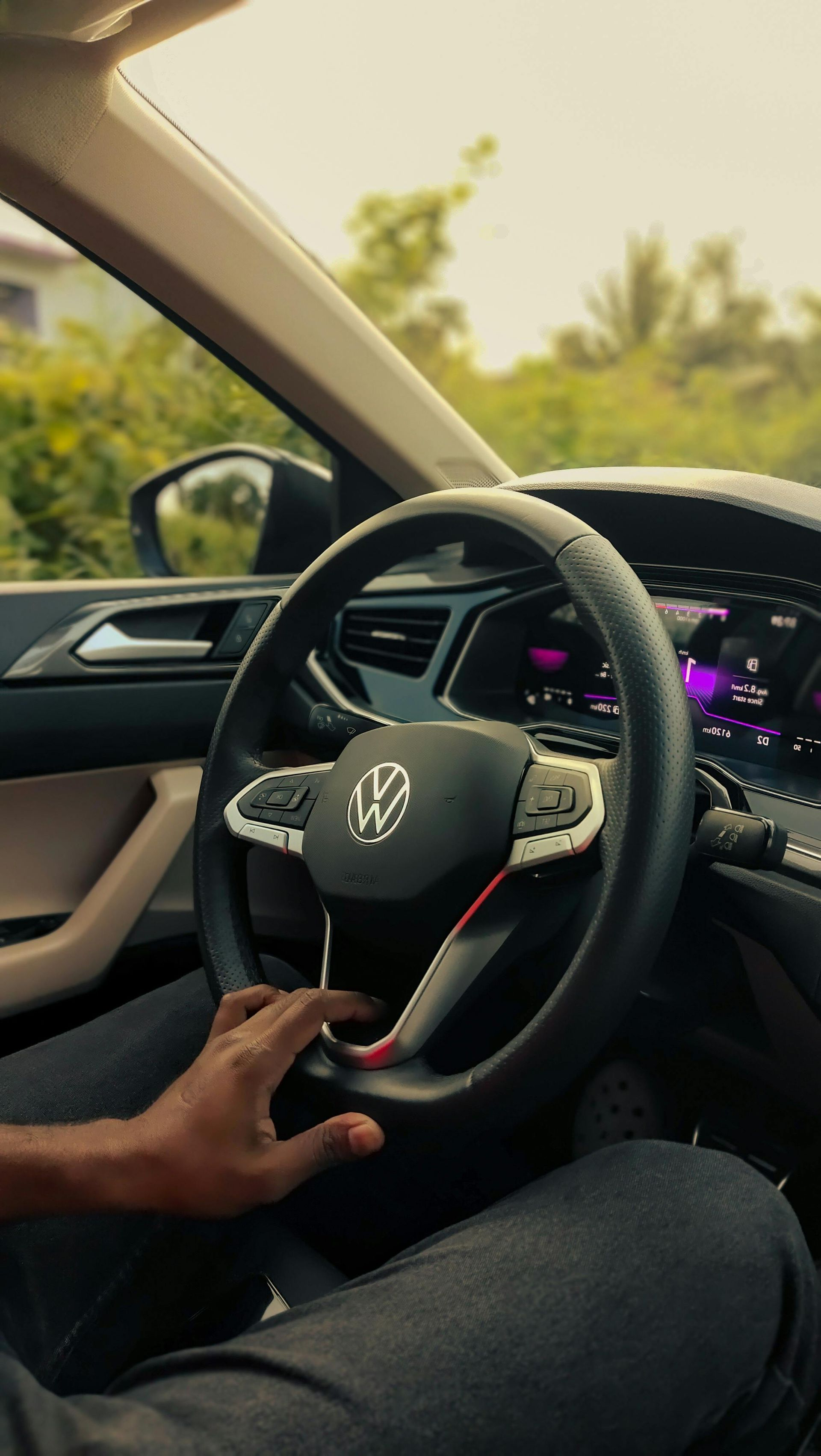 Hand on steering wheel inside a Volkswagen car, with dashboard lights glowing and trees visible outside.