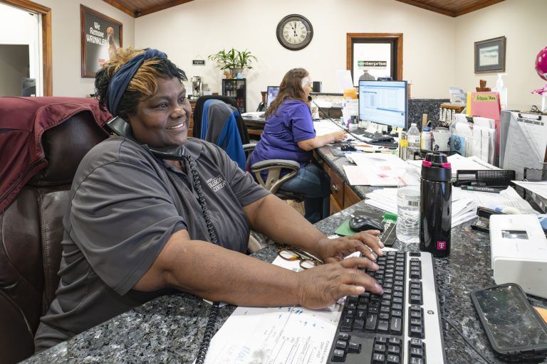 Person typing at a desk in a busy office with computer monitors and paperwork.