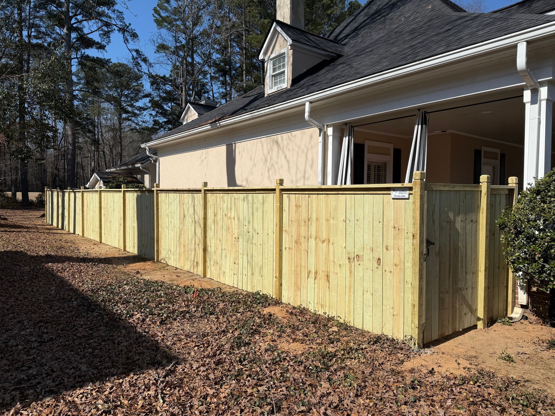 Wooden privacy fence along the side of a beige house with a porch in a sunny yard.