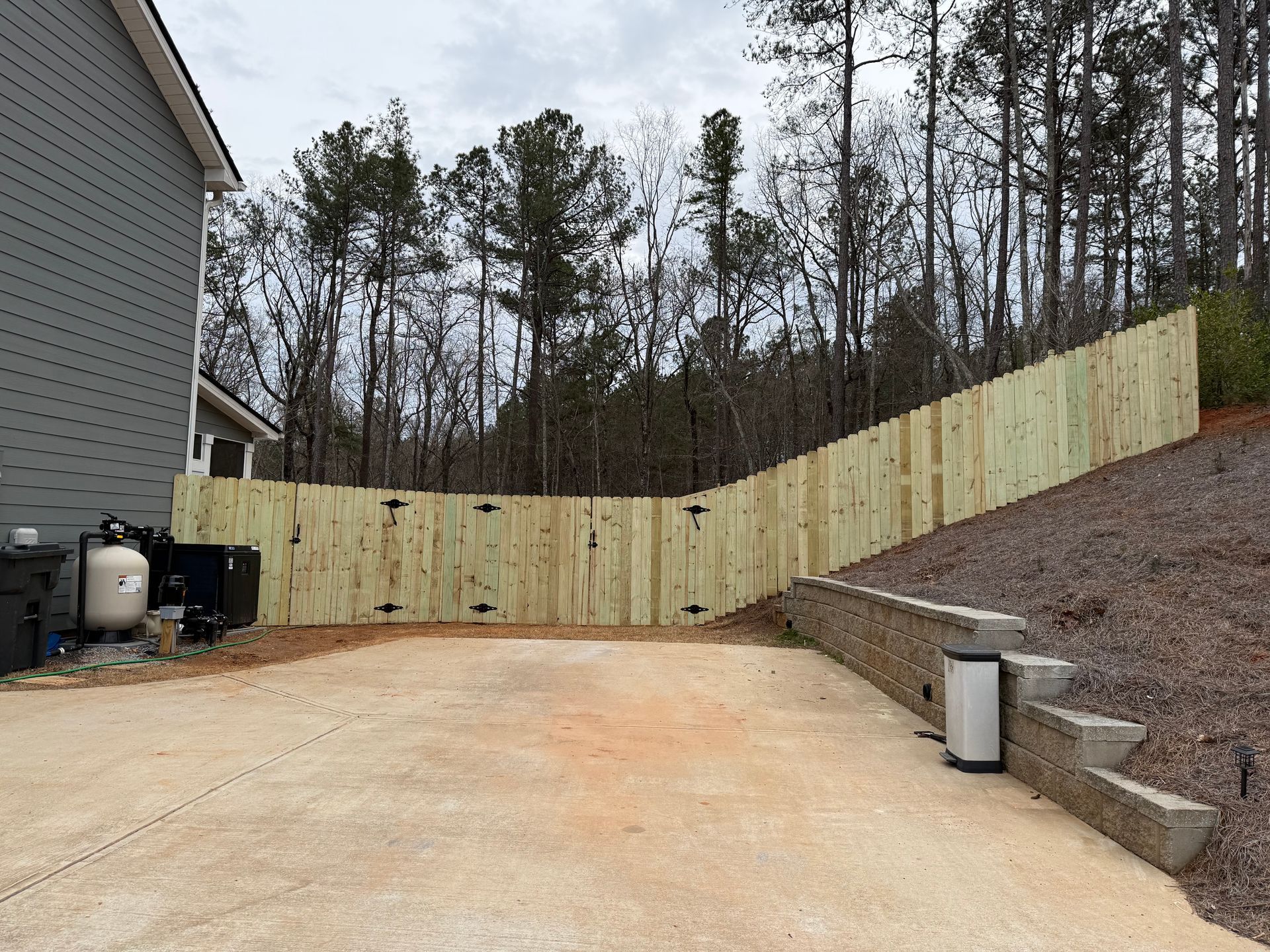 Concrete driveway beside house and retaining wall, bordered by a sloped wooded yard and tall pine trees.