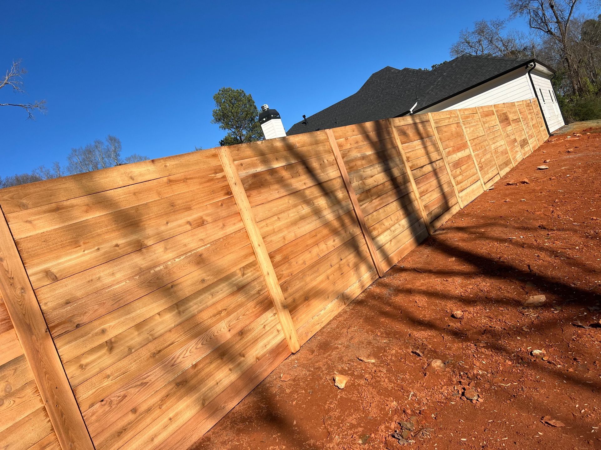 Wooden privacy fence along a sloped dirt yard beside a house under a clear blue sky.
