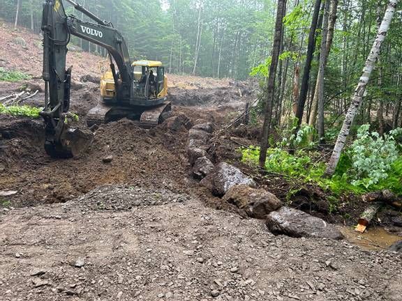 Une pelleteuse creuse dans un sol boueux près d'une zone boisée. Des pierres sont entassées en bordure.