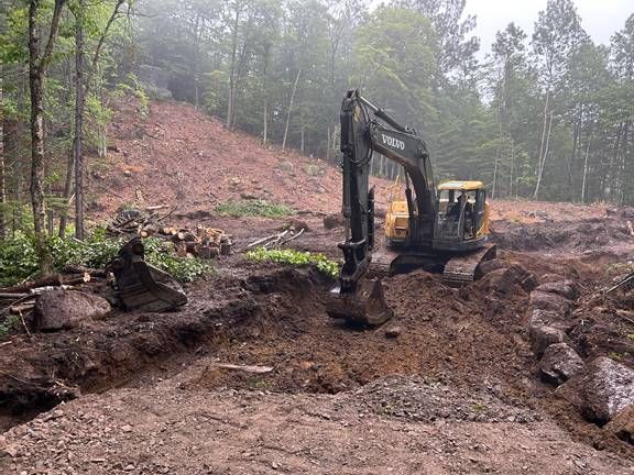 Une pelleteuse creuse dans un chantier boueux, des arbres en arrière-plan, ciel couvert.