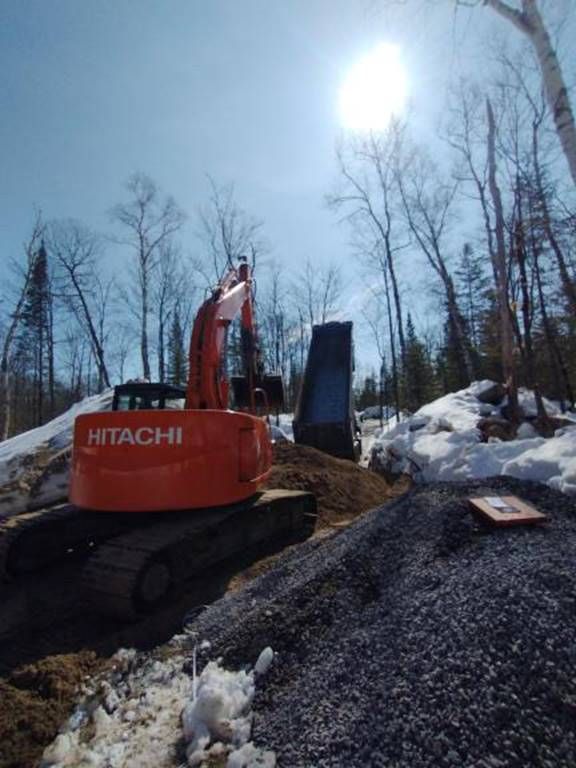 Une pelle mécanique Hitachi orange charge du gravier dans un camion-benne bleu par une journée ensoleillée.