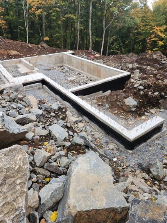 Fondations en béton pour un bâtiment en construction, entourées de rochers et de terre. Application d'une membrane d'étanchéité noire.
