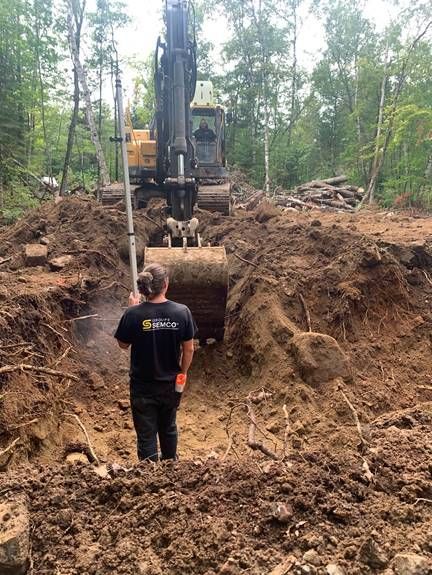 Un homme utilise un niveau pour mesurer une tranchée creusée par une excavatrice dans une zone boisée.