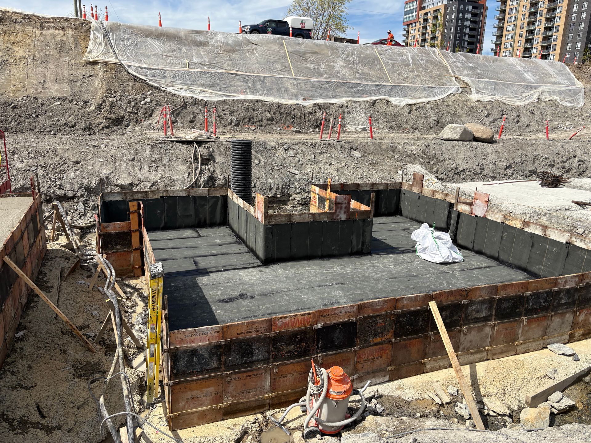 Chantier de construction ; coffrage de fondation dans une fosse excavée, mur de soutènement avec filet en arrière-plan.