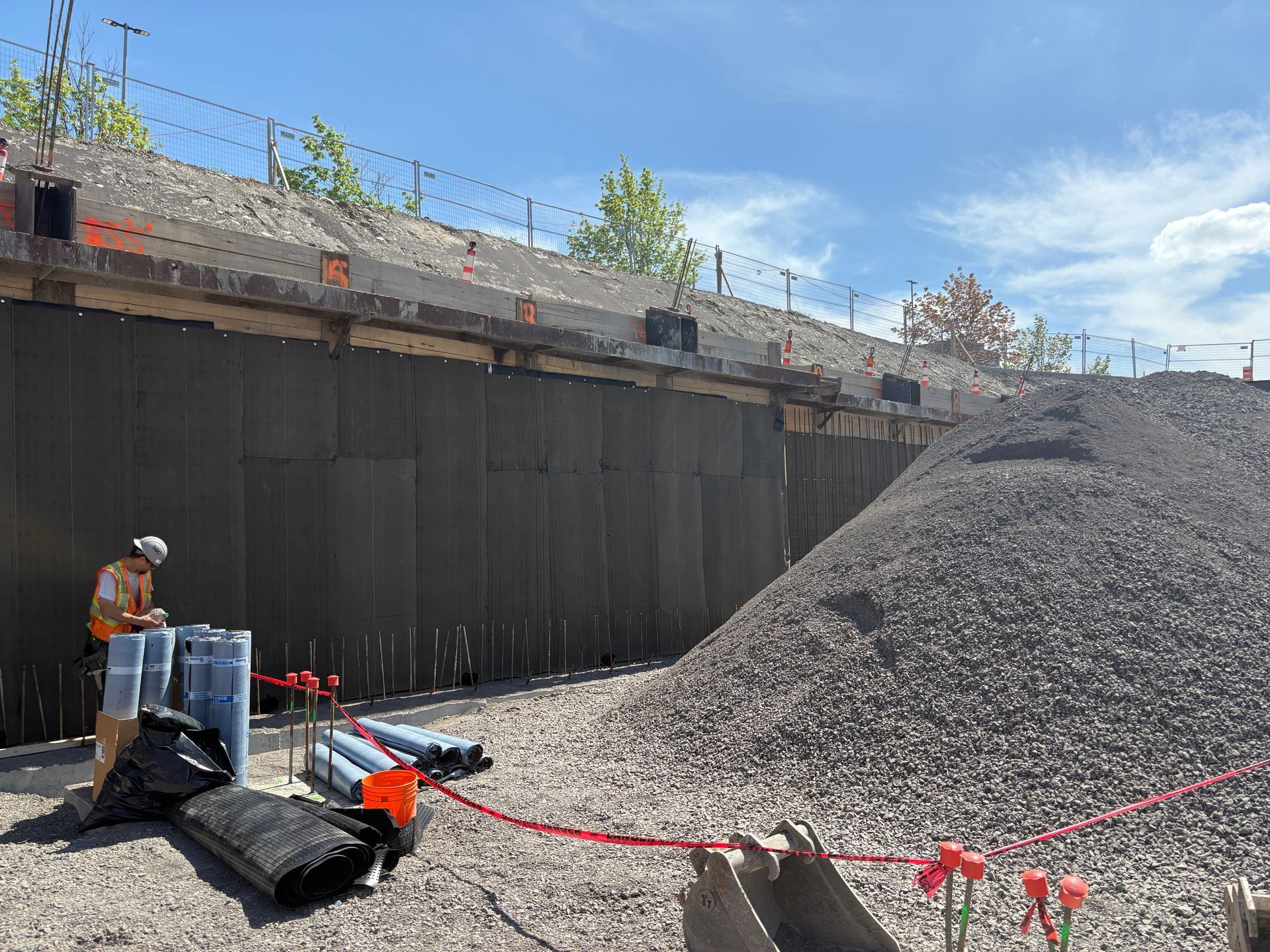 Chantier de construction avec un ouvrier ; un mur recouvert de matériau noir, un tas de gravier et des tuyaux bleus.