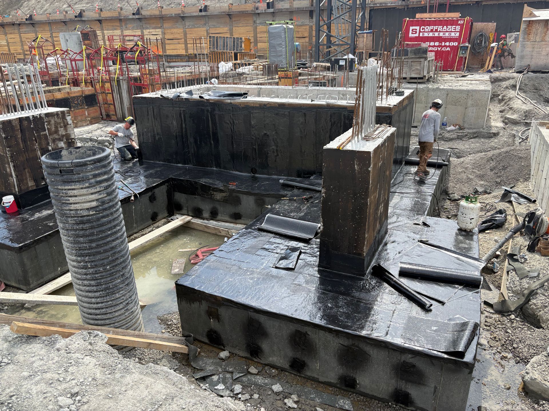 Chantier de construction avec fondations en béton, recouvertes d'un enduit noir. Des ouvriers sont présents.