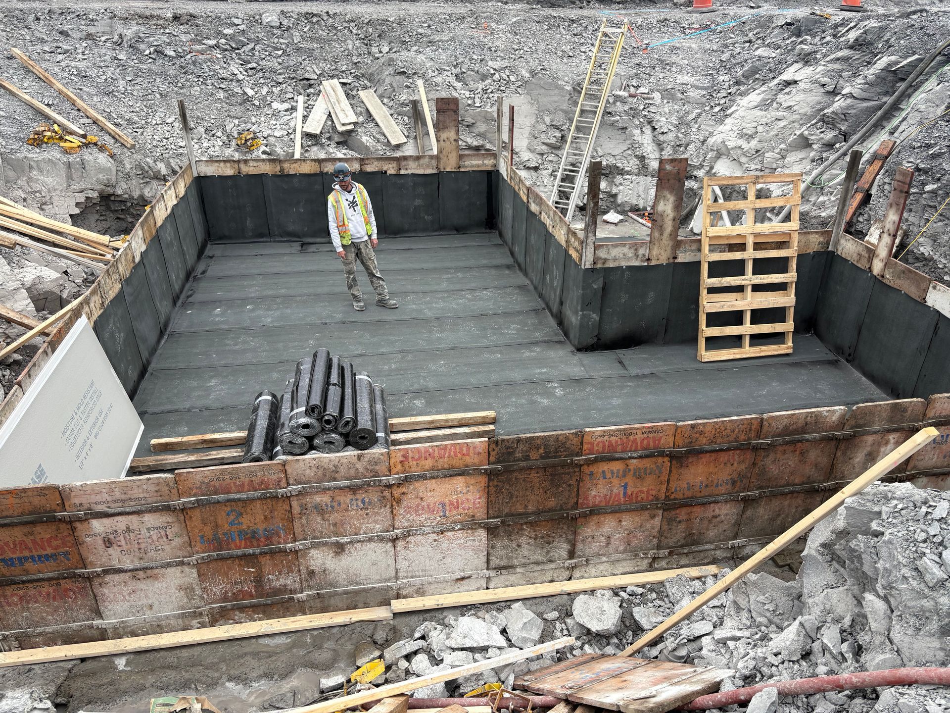 Chantier de construction : Structure de fondation en béton avec un ouvrier à l'intérieur ; revêtement noir.