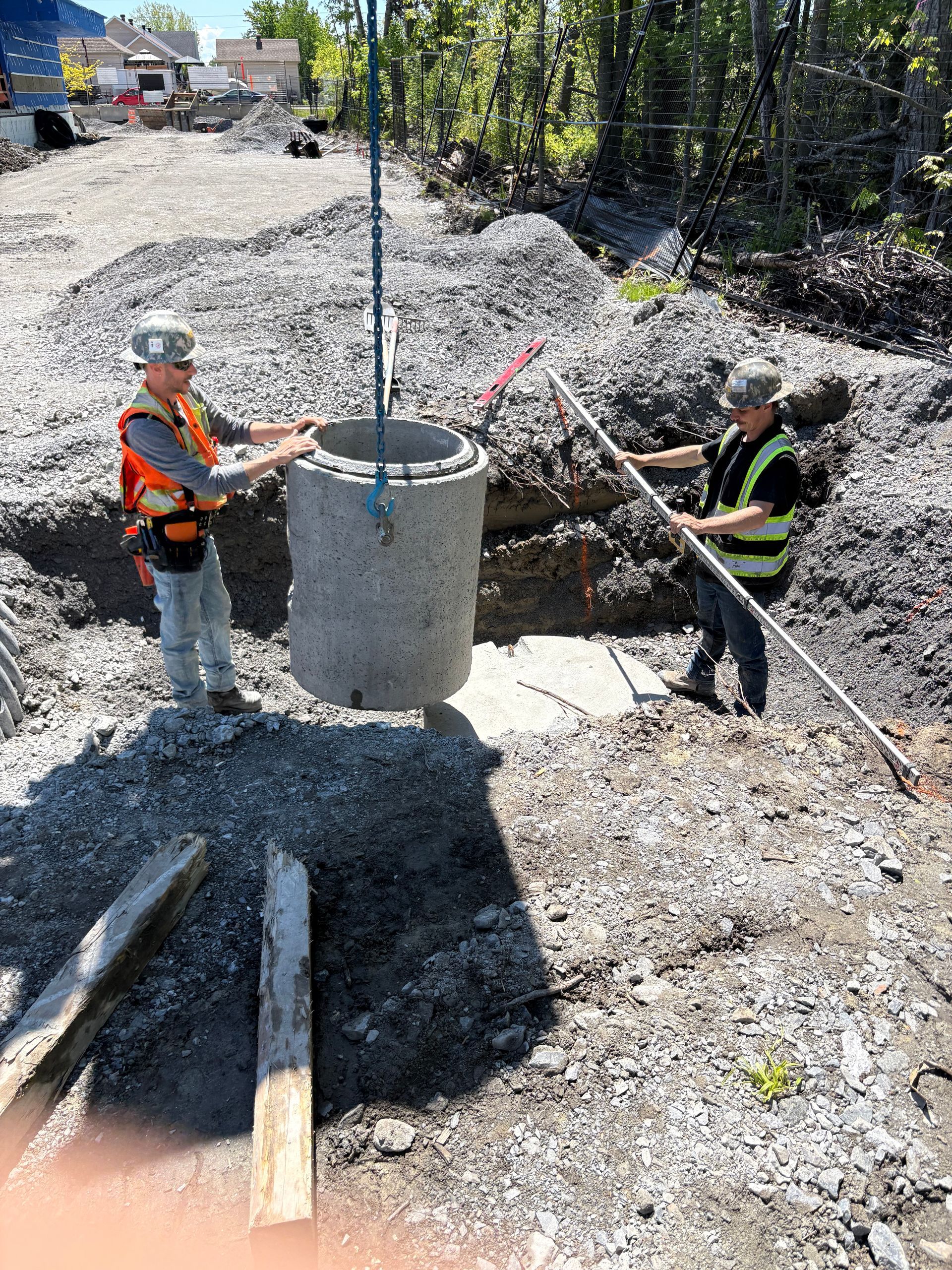 Deux ouvriers soulèvent un anneau d'égout en béton. L'un d'eux utilise une règle graduée. Chantier.