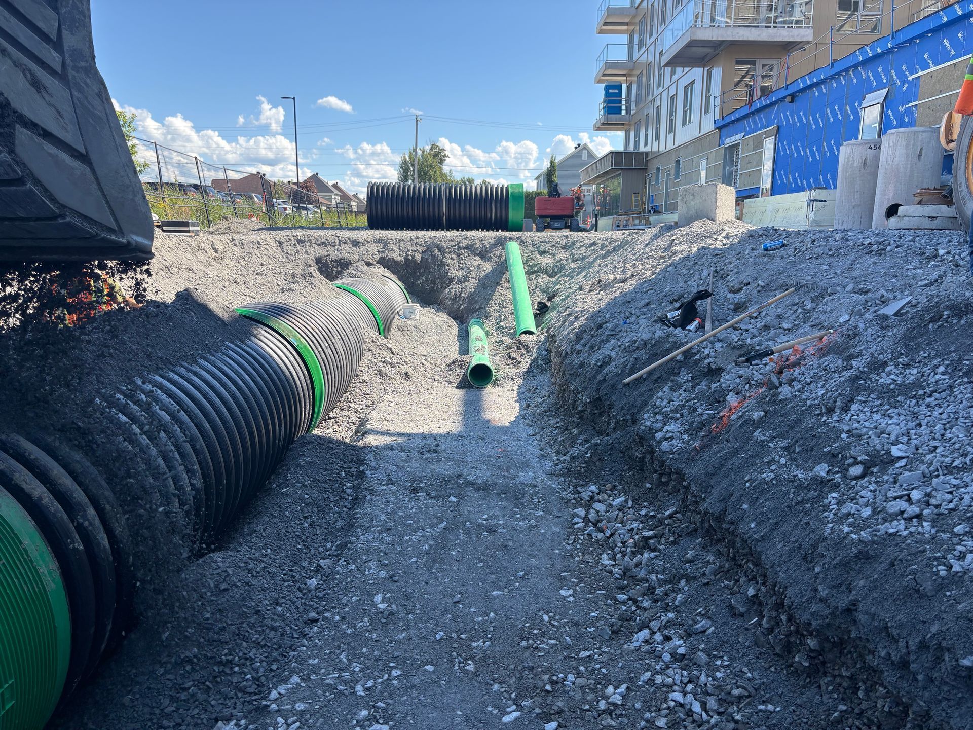 Chantier de construction avec des tuyaux verts et noirs dans une tranchée, à côté d'un bâtiment.