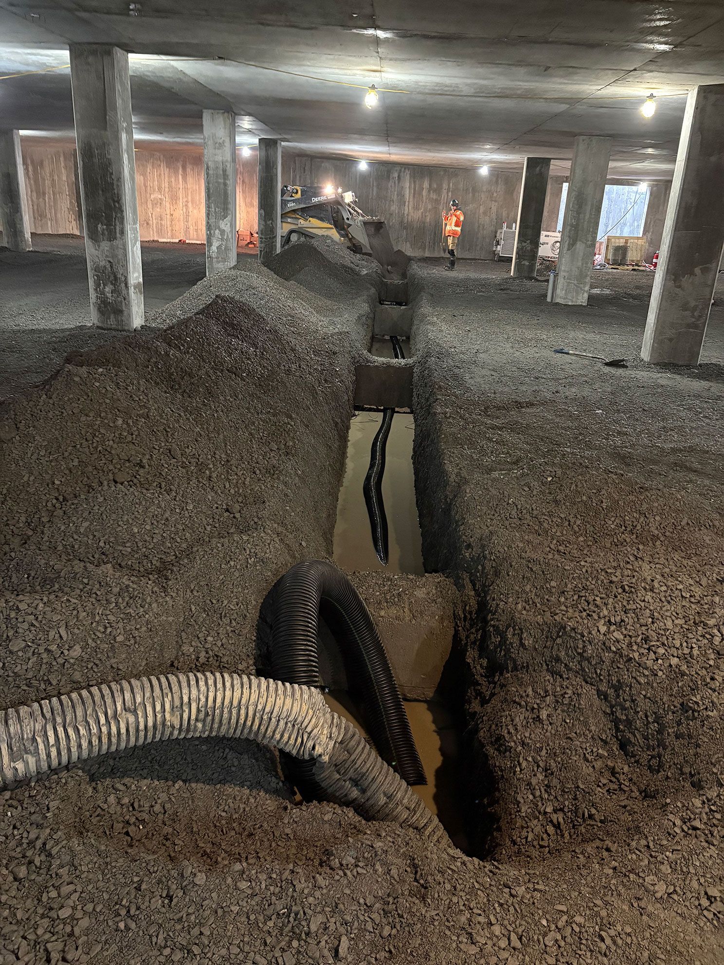 Chantier de construction avec une tranchée creusée dans du gravier ; tuyaux noirs et un ouvrier en arrière-plan.