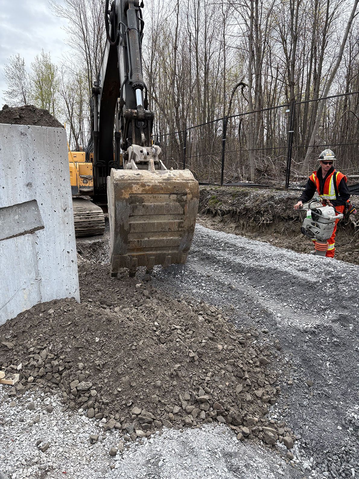 Une excavatrice creuse dans une zone de gravier à côté d'une structure en béton ; un ouvrier à proximité manœuvre l'engin.