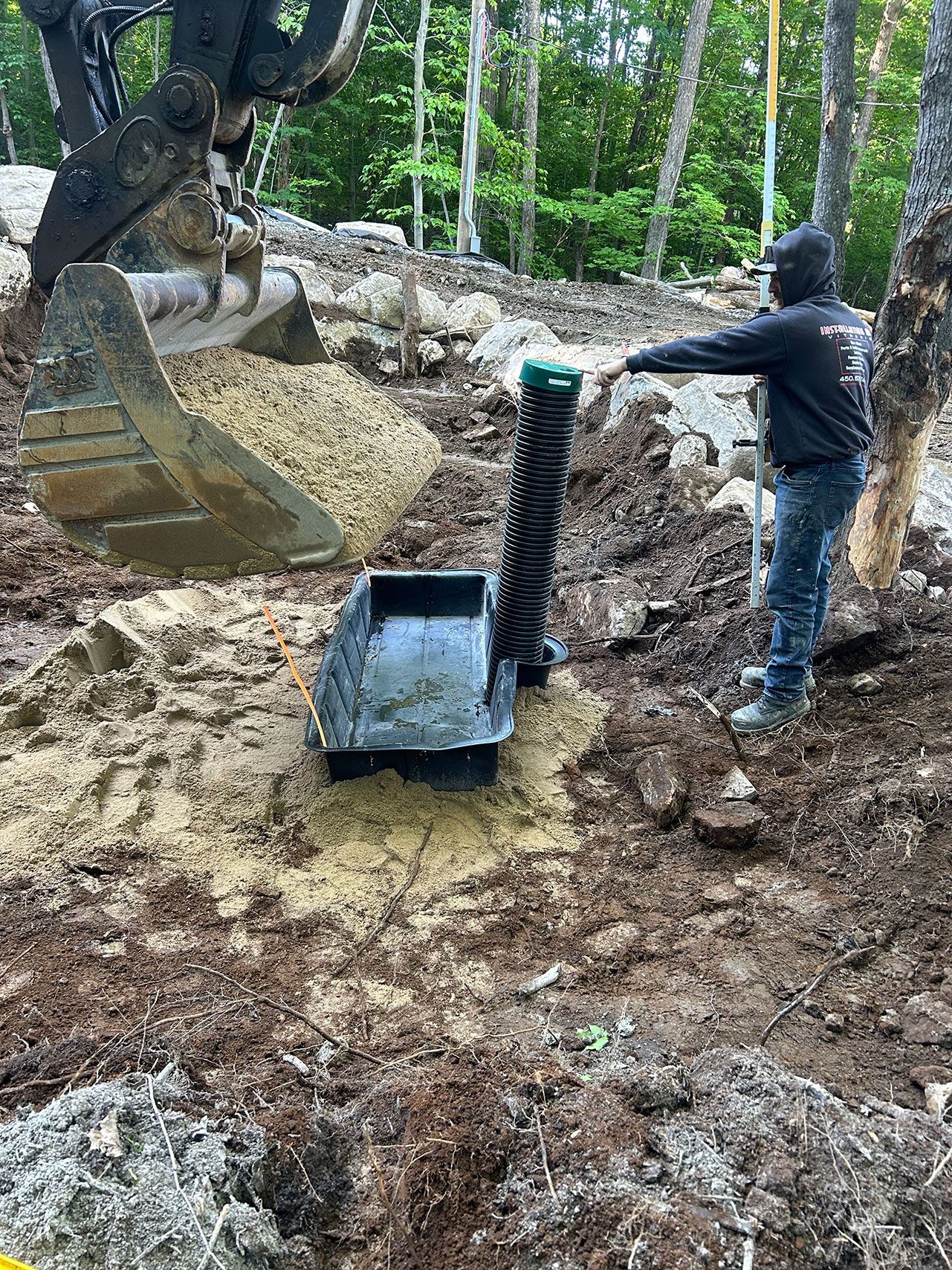 Une personne se trouve près d'une fosse septique excavée, le godet d'une excavatrice est à proximité. En extérieur, des arbres sont visibles.