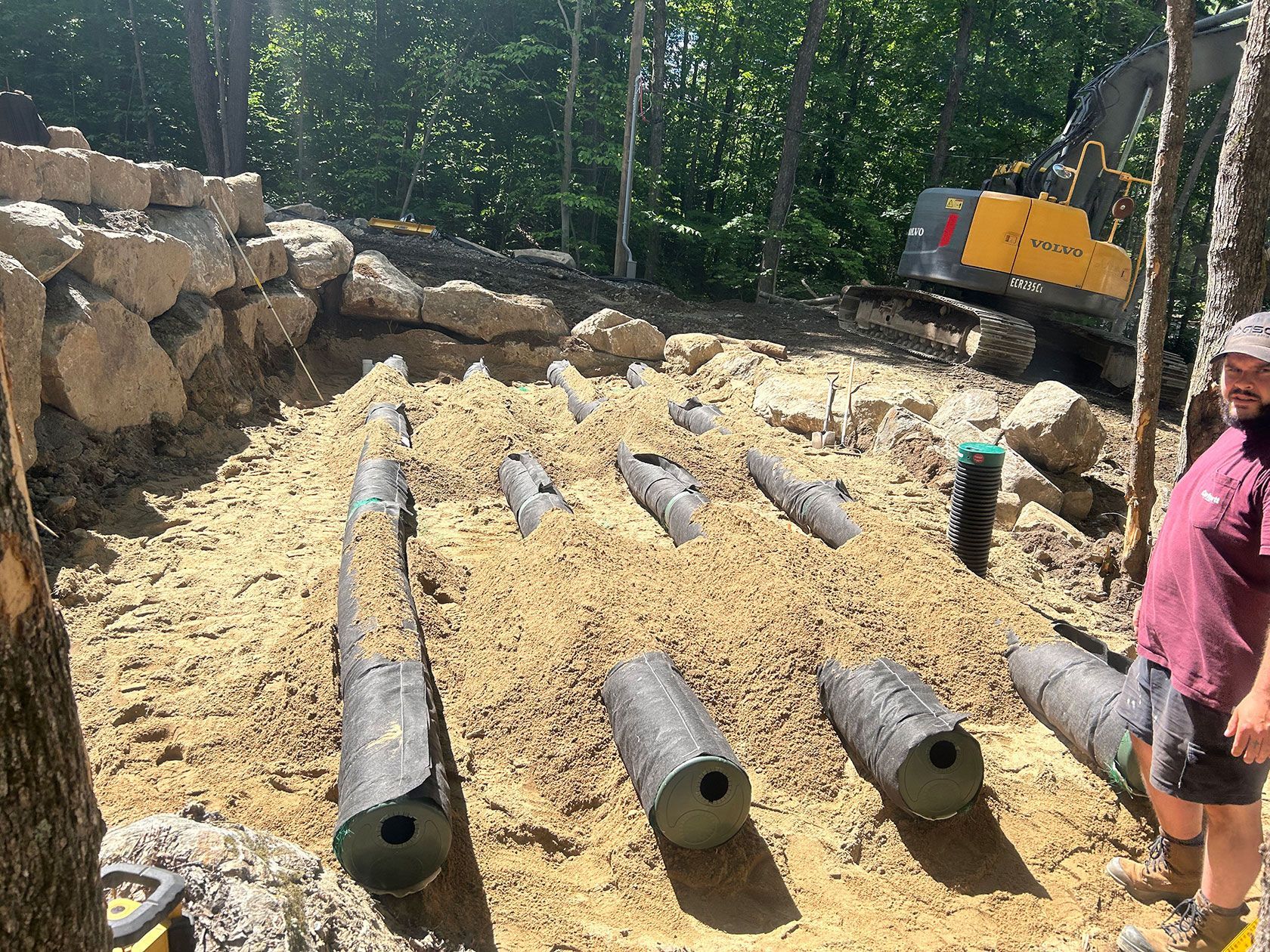 Chantier de construction avec terrain excavé, canalisations enterrées et un homme observant près des arbres ; une excavatrice est visible.