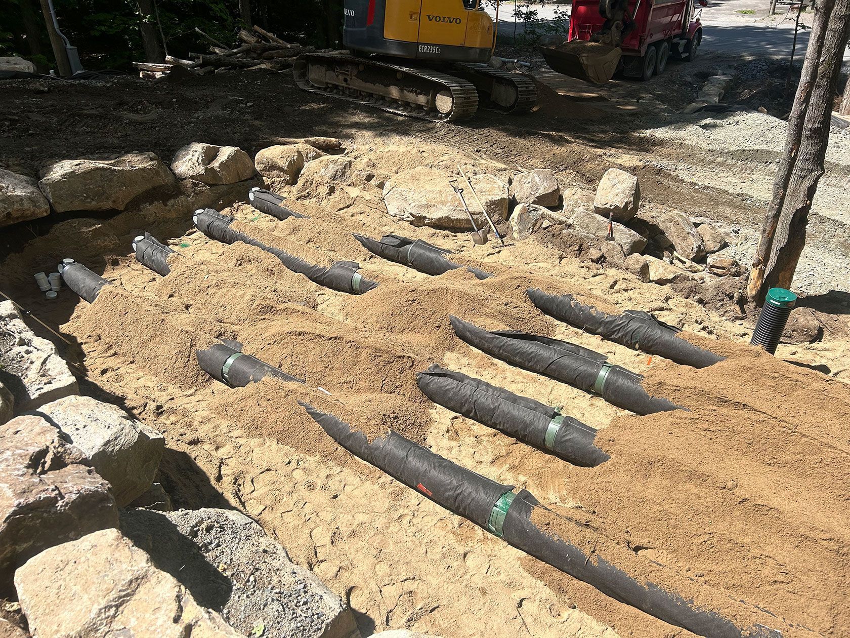 Chantier de construction avec des tuyaux noirs posés dans un sol sableux, le long d'un mur de soutènement en pierre et de machines.