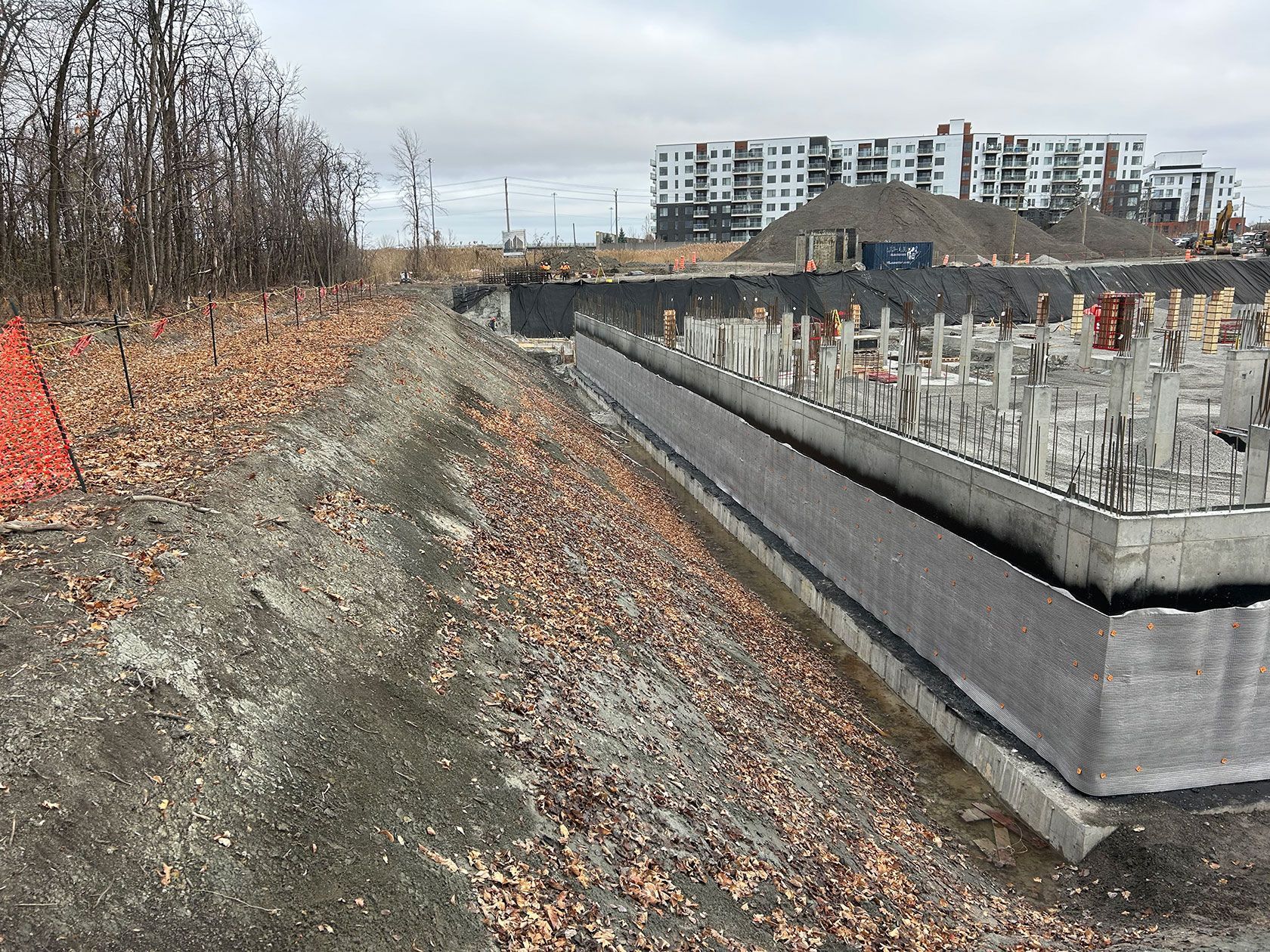 Chantier de construction avec fondations en béton, terre et feuilles mortes. Grand immeuble d'appartements en arrière-plan.
